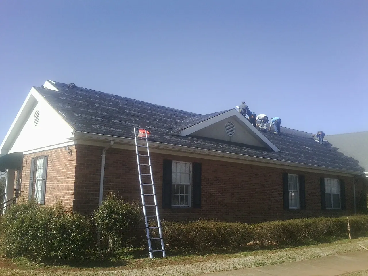 Crew tearing shingles off a brick ranch roof with a ladder set — Metal Roof Repair work by our crew in Daleville